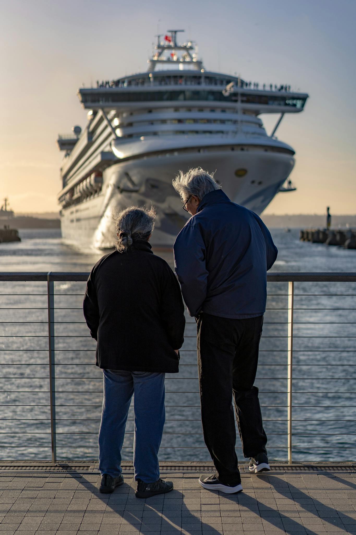 Senior couple standing at railing, admiring large cruise ship approaching dock at sunset.